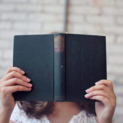 A woman holds an open book, reading in front of a brick wall.